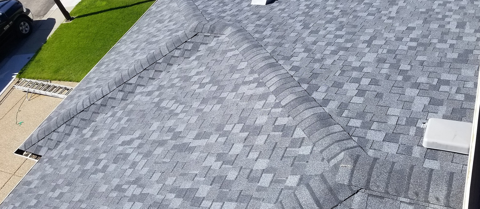 An aerial view of a gray asphalt shingle roof with visible ridges and a white vent.