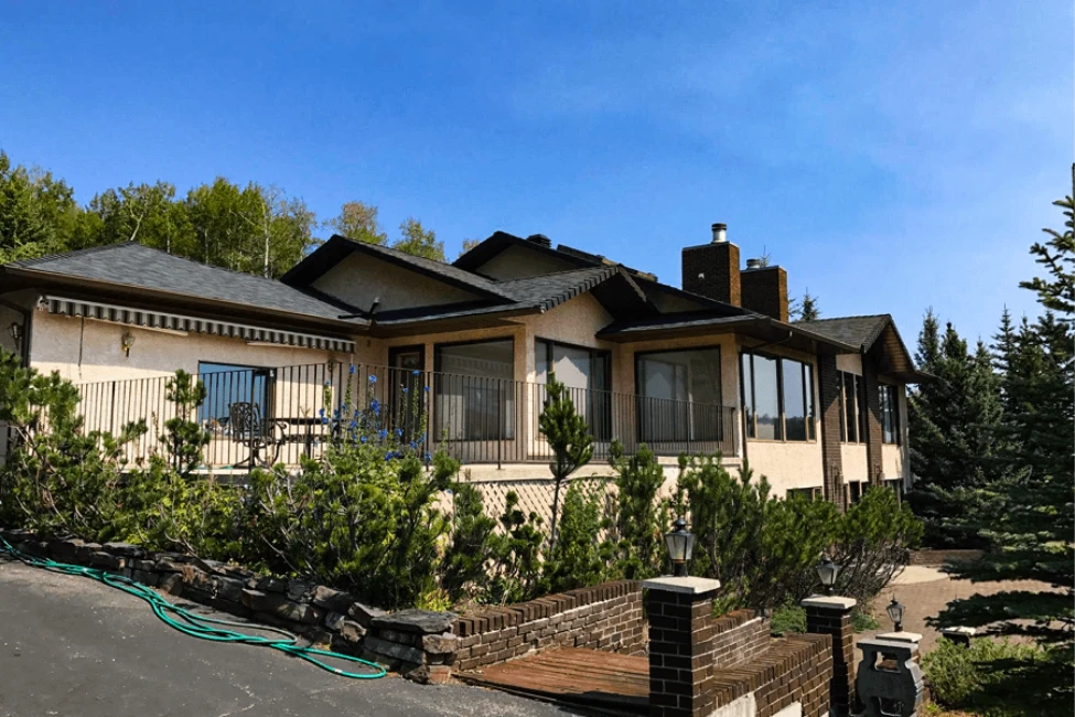 A beige two-story house with a balcony and surrounding greenery under a clear blue sky.
