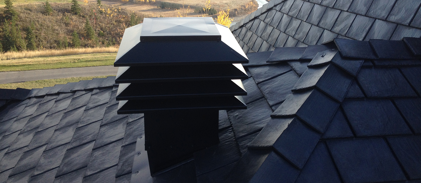 A black multi-tiered roof vent sits atop dark shingles with a scenic autumn landscape in the background.