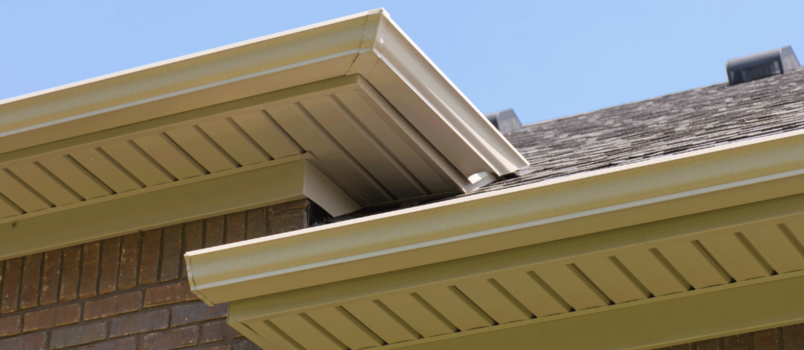 Close-up of beige gutters and siding on a brick house under a clear blue sky.
