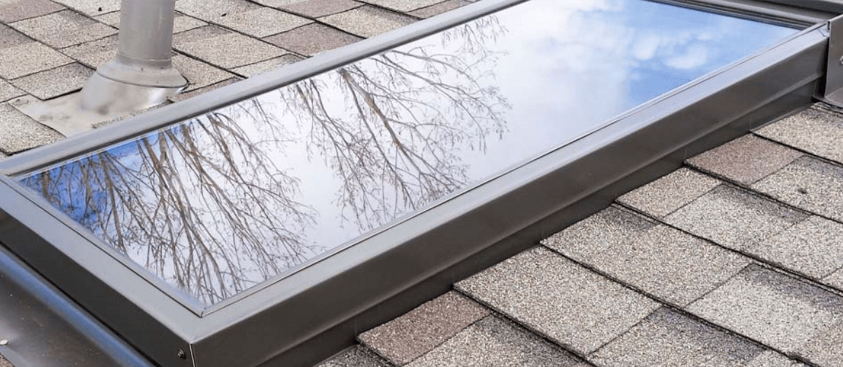 A rectangular skylight on a shingled roof reflects bare tree branches against a cloudy sky.