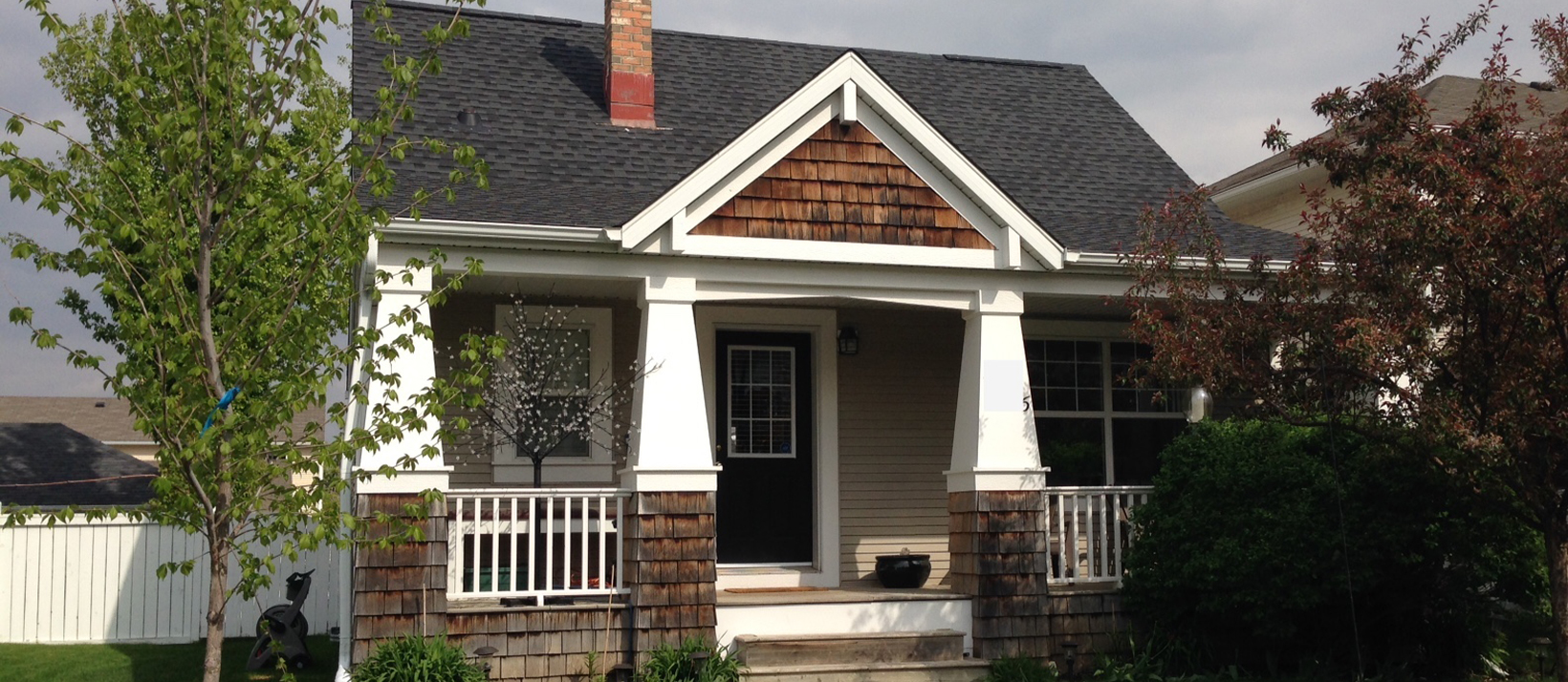 A charming single-story house with a white-columned porch and dark shingle roof, surrounded by greenery under an overcast sky.